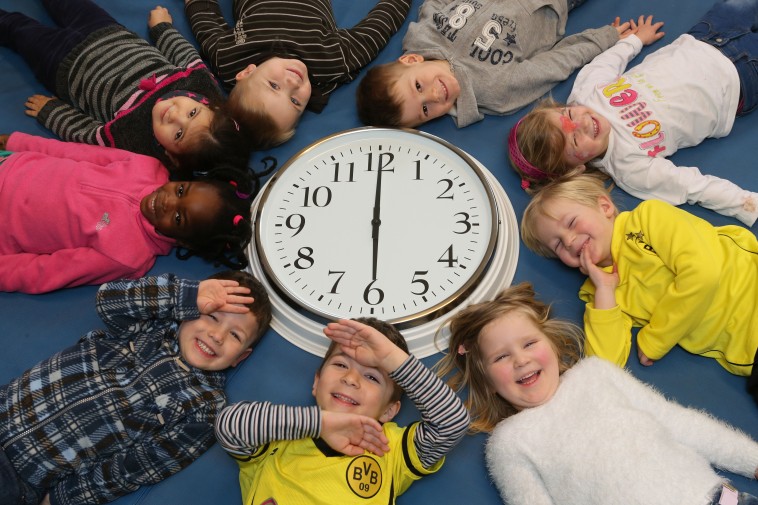 Kinder bilden liegend einen Kreis um eine Uhr herum.